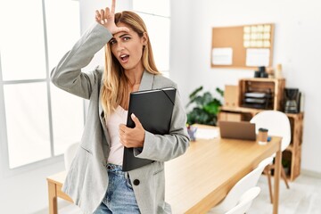 Blonde business woman at the office making fun of people with fingers on forehead doing loser gesture mocking and insulting.