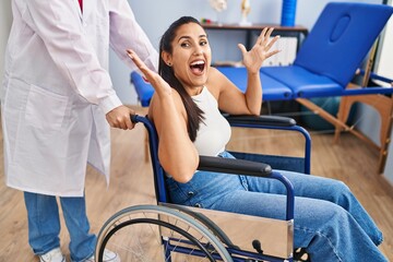 Obraz premium Young hispanic woman sitting on wheelchair at physiotherapy clinic celebrating victory with happy smile and winner expression with raised hands
