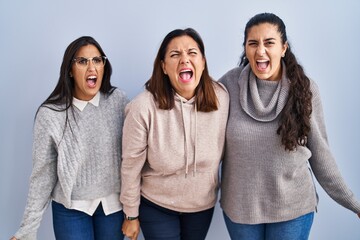 Mother and two daughters standing over blue background angry and mad screaming frustrated and furious, shouting with anger. rage and aggressive concept.