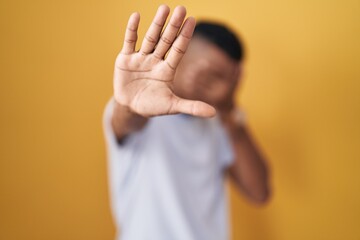 Young hispanic man standing over yellow background covering eyes with hands and doing stop gesture...