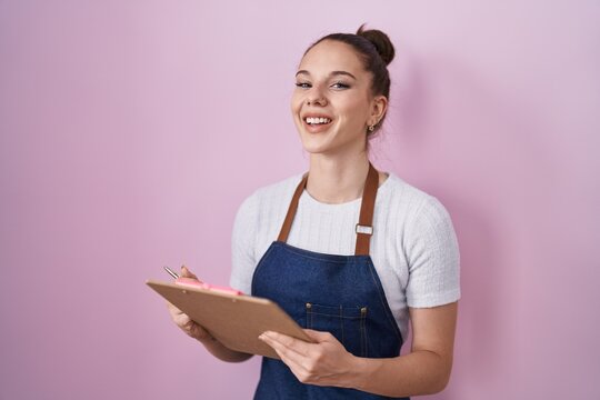 Young hispanic girl wearing professional waitress apron taking order smiling and laughing hard out loud because funny crazy joke.