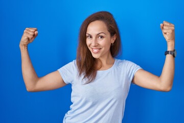 Fototapeta premium Brunette woman standing over blue background showing arms muscles smiling proud. fitness concept.