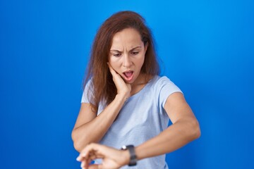 Brunette woman standing over blue background looking at the watch time worried, afraid of getting late