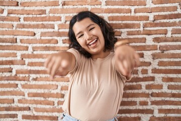 Young hispanic woman standing over bricks wall pointing to you and the camera with fingers, smiling positive and cheerful