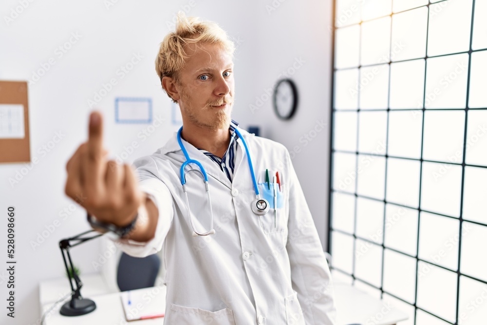 Sticker Young blond man wearing doctor uniform and stethoscope at clinic showing middle finger, impolite and rude fuck off expression