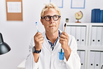 Young blond dentist man working at dentist clinic holding toothbrushes smiling looking to the side and staring away thinking.