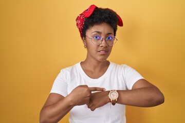 Young african american woman standing over yellow background in hurry pointing to watch time,...