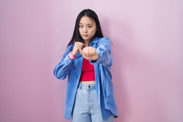 Young asian woman standing over pink background punching fist to fight, aggressive and angry...