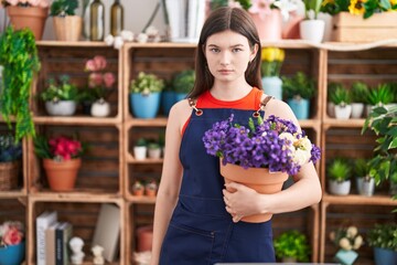 Young caucasian woman working at florist shop holding pot with flowers thinking attitude and sober expression looking self confident