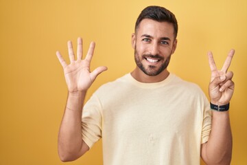 Handsome hispanic man standing over yellow background showing and pointing up with fingers number seven while smiling confident and happy.