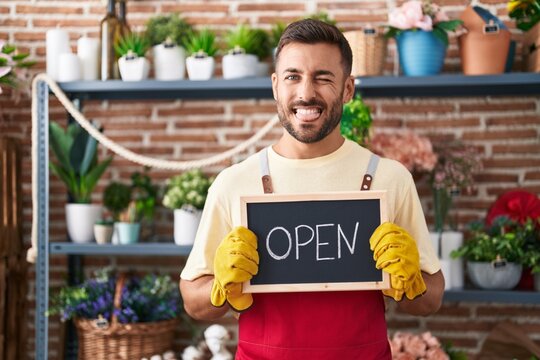 Handsome hispanic man working at florist holding open sign sticking tongue out happy with funny expression.
