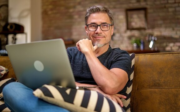 Happy middle aged man sitting on couch at home working with laptop computer, smiling.