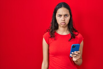 Young brazilian woman using smartphone over red background skeptic and nervous, frowning upset because of problem. negative person.
