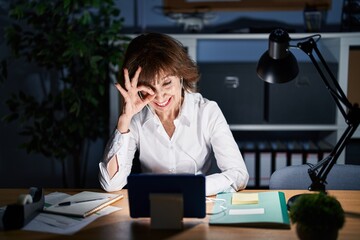 Middle age woman working at the office at night doing ok gesture with hand smiling, eye looking through fingers with happy face.