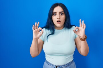 Young modern girl with blue hair standing over blue background looking surprised and shocked doing ok approval symbol with fingers. crazy expression