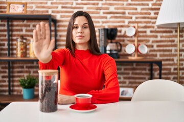 Young hispanic woman drinking coffee at home with open hand doing stop sign with serious and confident expression, defense gesture