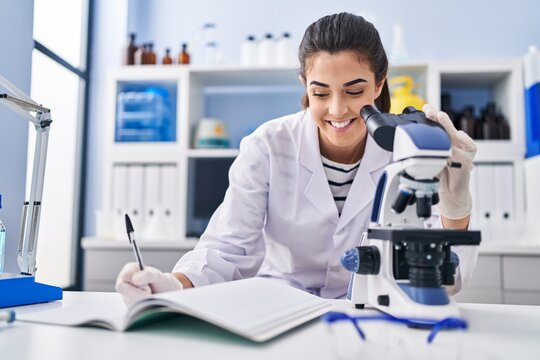 Young beautiful hispanic woman scientist using microscope writing on notebook at laboratory