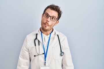 Young hispanic man wearing doctor uniform and stethoscope making fish face with lips, crazy and comical gesture. funny expression.