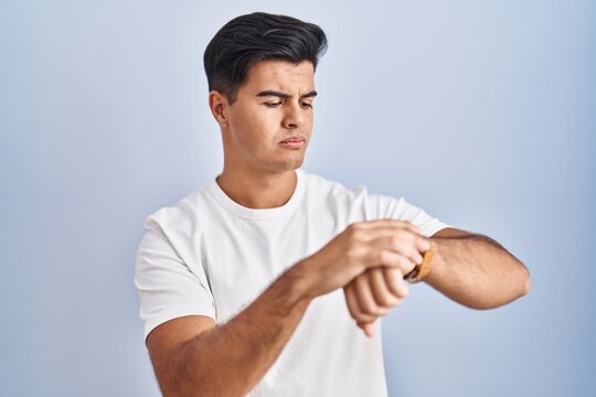 Hispanic man standing over blue background checking the time on wrist watch, relaxed and confident