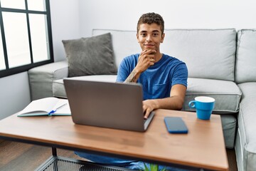 Young handsome hispanic man using laptop sitting on the floor looking confident at the camera with smile with crossed arms and hand raised on chin. thinking positive.