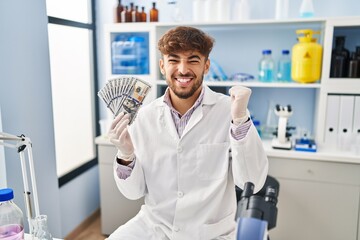 Arab man with beard working at scientist laboratory holding money screaming proud, celebrating victory and success very excited with raised arms