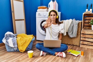 Young hispanic woman studying while waiting for laundry doing stop sing with palm of the hand. warning expression with negative and serious gesture on the face.