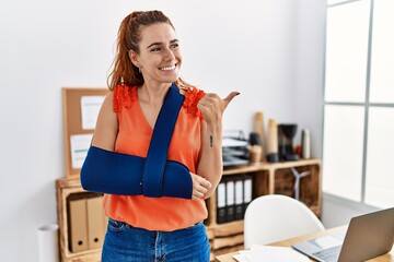 Young redhead woman wearing arm on sling at the office pointing thumb up to the side smiling happy with open mouth