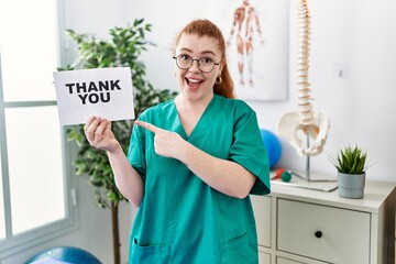 Young redhead physiotherapist woman working at pain recovery clinic holding thank you banner...