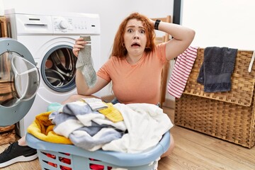 Young redhead woman putting dirty laundry into washing machine crazy and scared with hands on head,...
