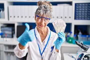 Middle age woman working at scientist laboratory holding dollars smiling happy and positive, thumb up doing excellent and approval sign