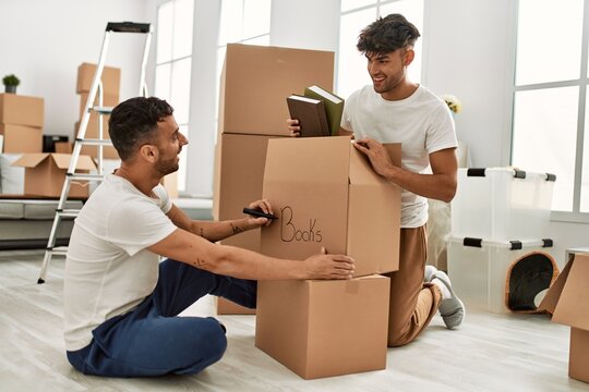 Two hispanic men couple smiling confident packing book cardboard box at new home
