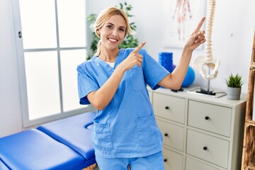 Beautiful blonde physiotherapist woman working at pain recovery clinic smiling and looking at the camera pointing with two hands and fingers to the side.