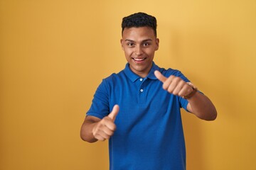 Young hispanic man standing over yellow background approving doing positive gesture with hand, thumbs up smiling and happy for success. winner gesture.