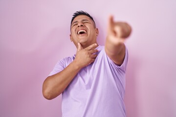 Young hispanic man standing over pink background laughing at you, pointing finger to the camera...