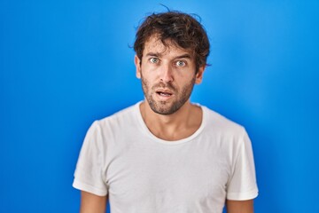 Hispanic young man standing over blue background in shock face, looking skeptical and sarcastic,...