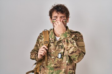 Hispanic young man wearing camouflage army uniform smelling something stinky and disgusting, intolerable smell, holding breath with fingers on nose. bad smell