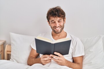 Young man reading book sitting on bed at bedroom