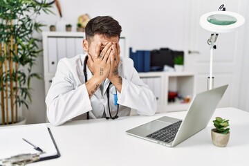 Young doctor working at the clinic using computer laptop with sad expression covering face with hands while crying. depression concept.