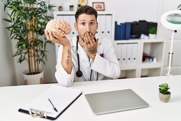 Young doctor holding brain at medical clinic covering mouth with hand, shocked and afraid for...