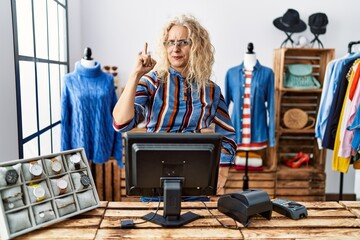 Middle age blonde woman working as manager at retail boutique pointing finger up with successful...