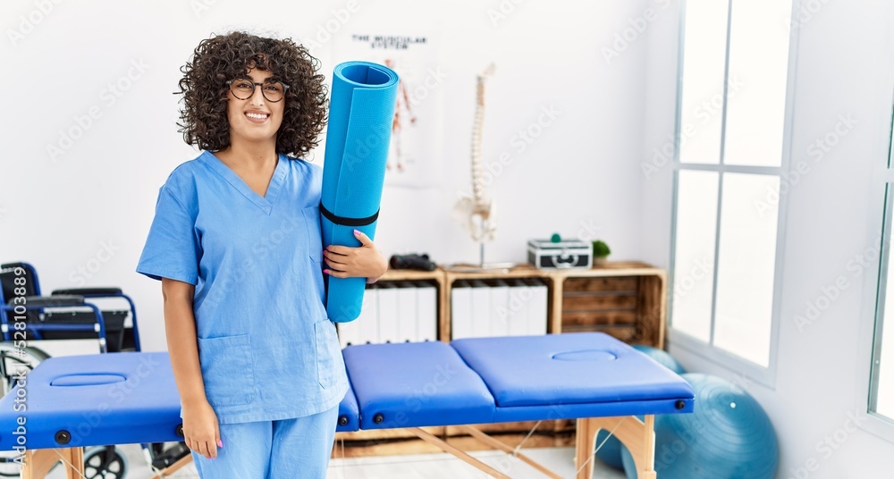 Wall mural young middle east woman wearing physio therapist uniform holding yoga mat at clinic