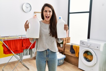 Young brunette woman holding detergent bottle at laundry room angry and mad screaming frustrated and furious, shouting with anger looking up.