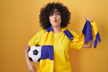 Young brunette woman with curly hair football hooligan holding ball making fish face with mouth and...