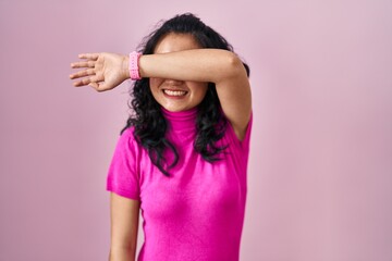 Young asian woman standing over pink background covering eyes with arm smiling cheerful and funny. blind concept.