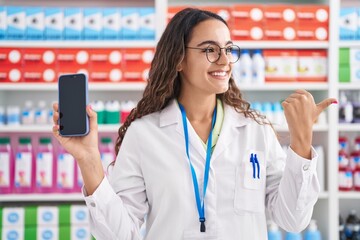 Young hispanic woman working at pharmacy drugstore showing smartphone screen winking looking at the camera with sexy expression, cheerful and happy face.