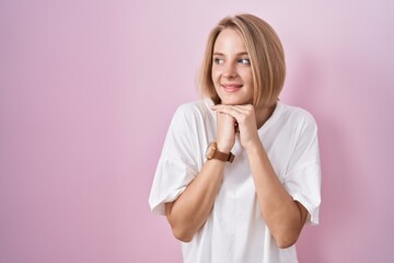 Young caucasian woman standing over pink background laughing nervous and excited with hands on chin looking to the side