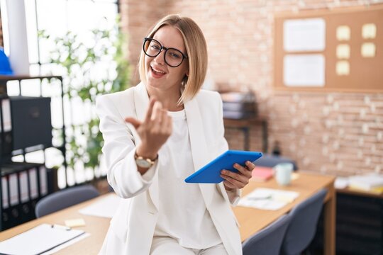 Young caucasian woman working at the office wearing glasses beckoning come here gesture with hand inviting welcoming happy and smiling