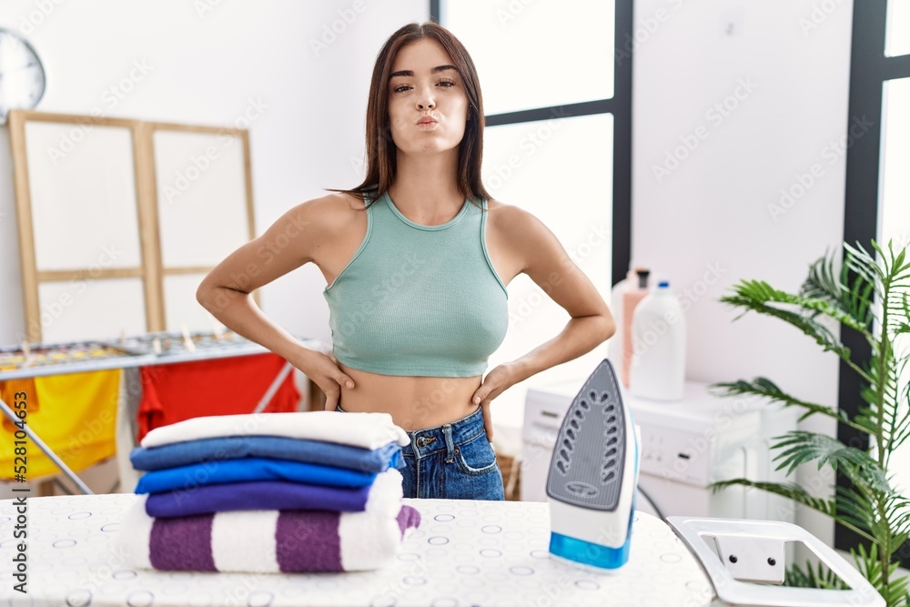 Poster Young hispanic woman ironing clothes at laundry room puffing cheeks with funny face. mouth inflated with air, crazy expression.