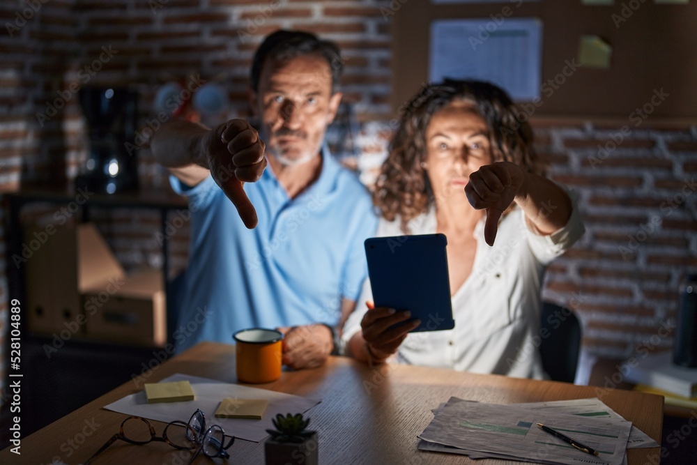 Sticker Middle age hispanic couple using touchpad sitting on the table at night looking unhappy and angry showing rejection and negative with thumbs down gesture. bad expression.