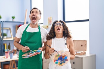 Middle age hispanic couple painting at art studio angry and mad screaming frustrated and furious,...